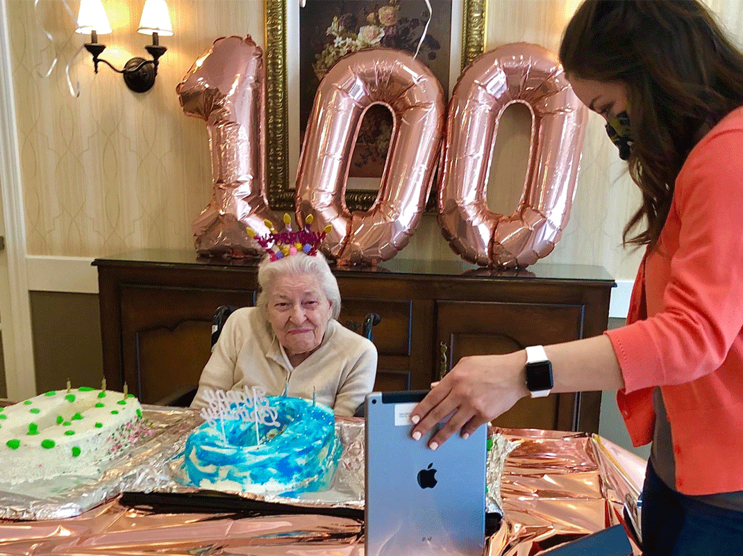 A woman celebrates her 100th birthday via Zoom with help from an assisted living center staff member holding a tablet.