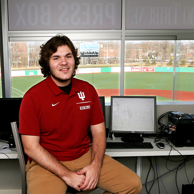 A man in a red IU polo shirt sits in a press box looking over a baseball field.