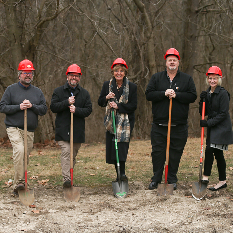 Five people with shovels and red construction hats break ground on a project.