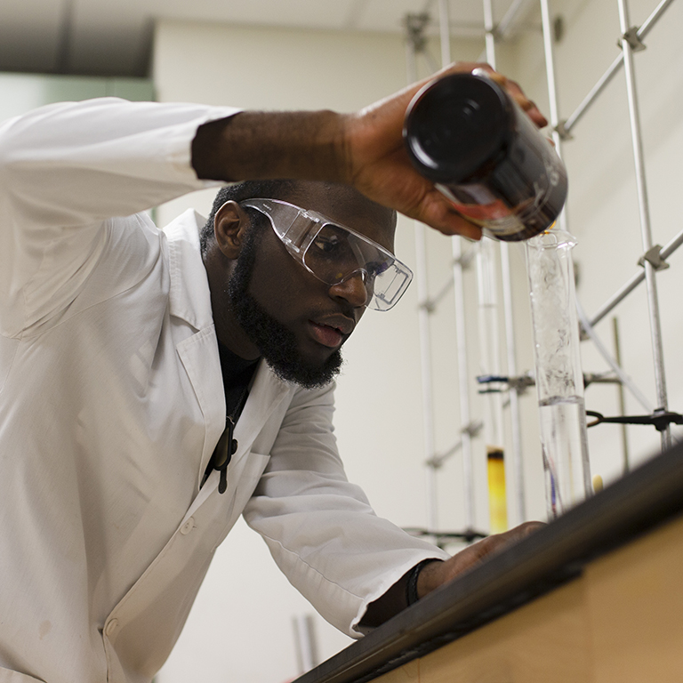 A man in a lab coat and glasses pours liquid into a container