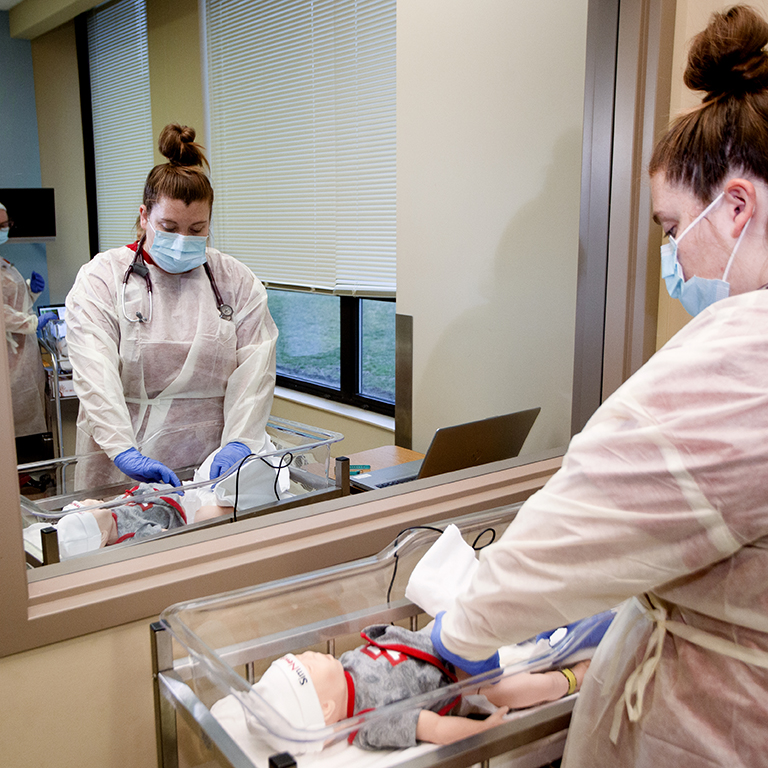 A woman in a mask and scrubs cares for an infant mannequin