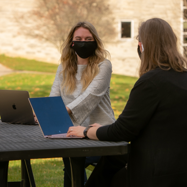 Two women in masks talk over laptops