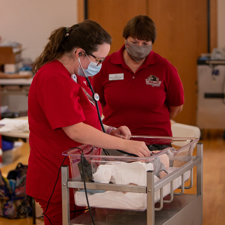 A woman in red scrubs uses a stethoscope on a baby mannequin while an instructor watches