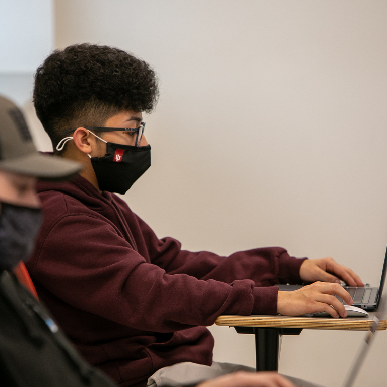 A man wearing a mask sits at a laptop