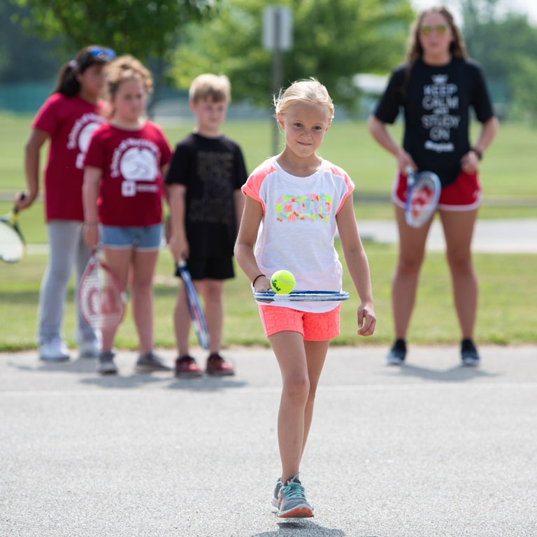 A child walks with a tennis racquet and ball.