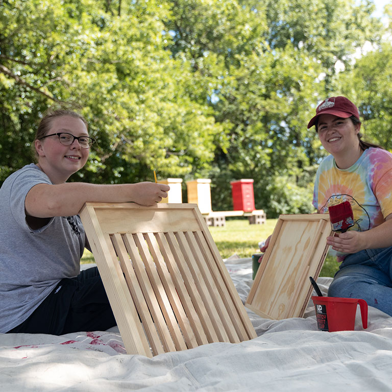 Two women pose for a picture while painting bee hives.
