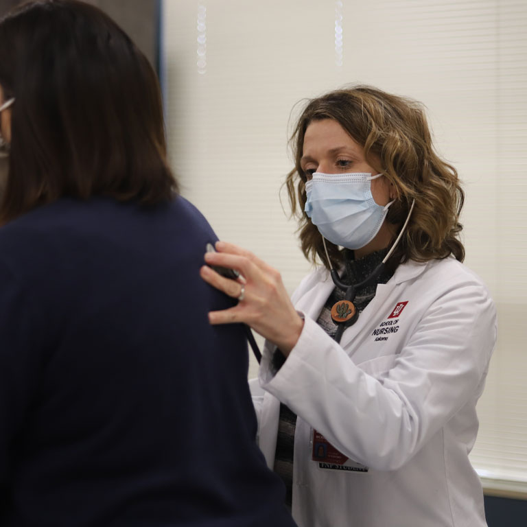 A woman in a white lab coat listens to a woman's heart with a stethoscope