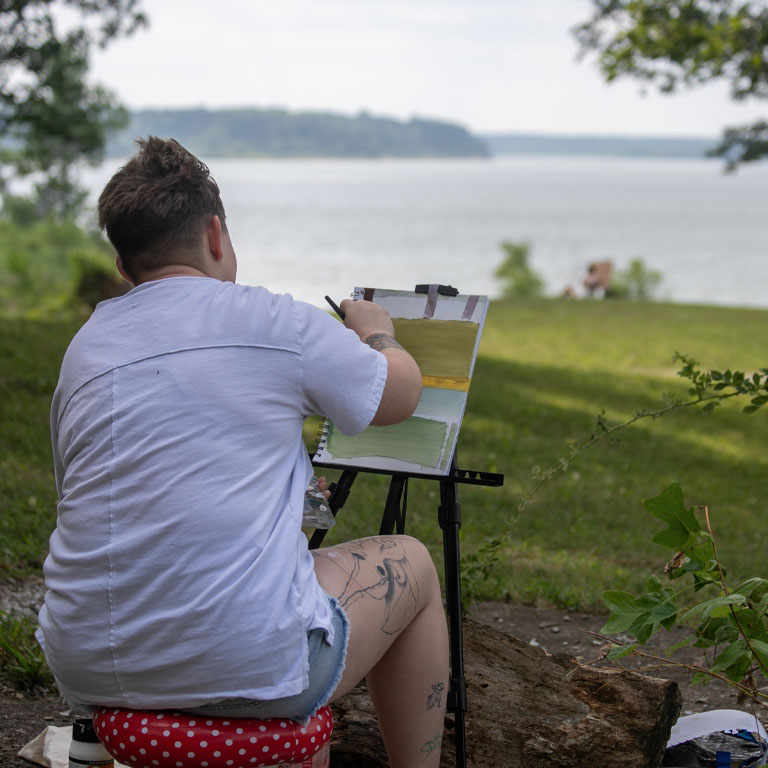 A student faces away from the camera, towards a reservoir, painting on a canvas.