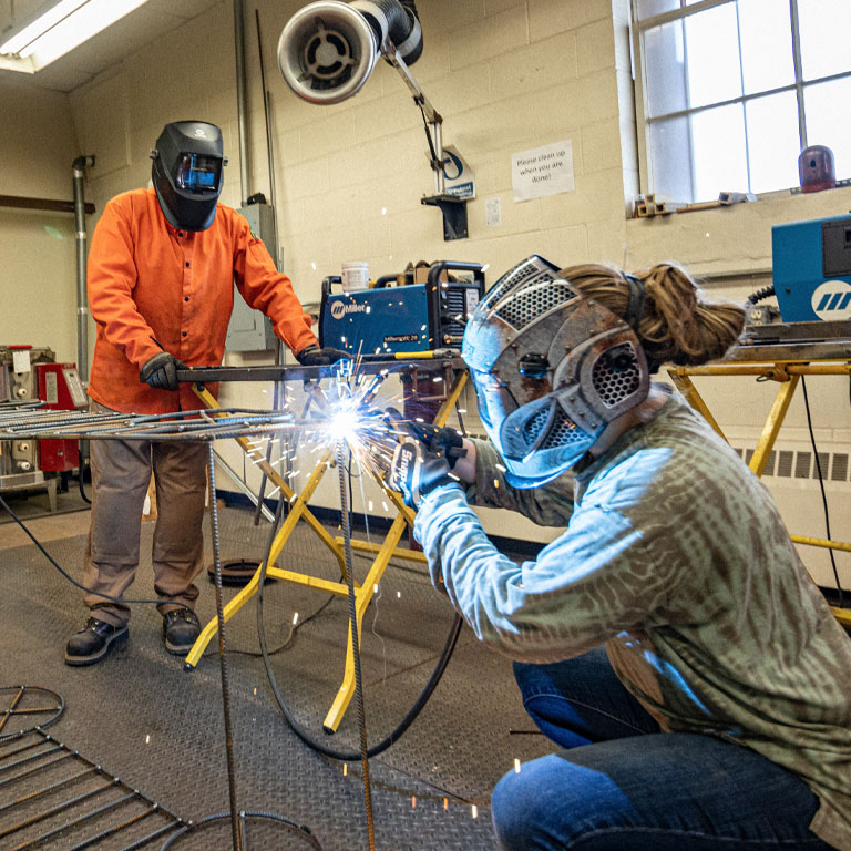 A student and faculty member in masks weld a metal car frame sculpture.