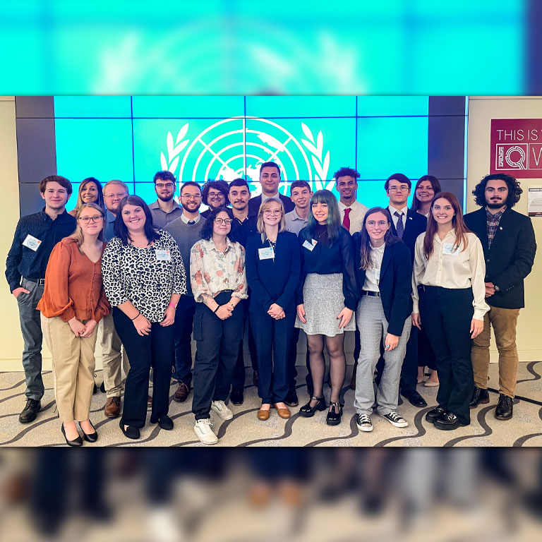 A large group of men and women stand in front of a Model UN logo on a wall.