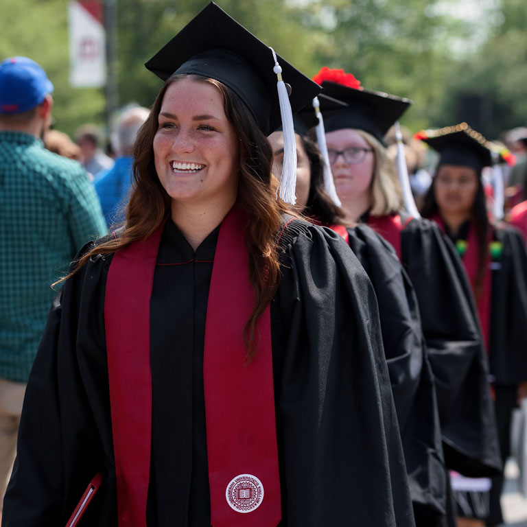 A woman wears a graduation cap and gown, with a line of people behind her.
