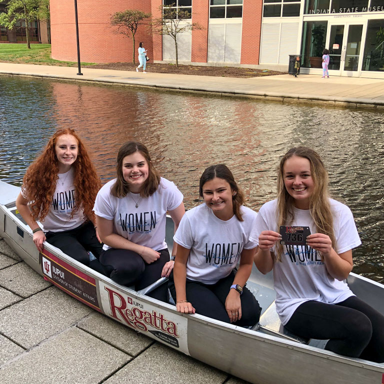 Four women pose in a steel canoe next to a canal.