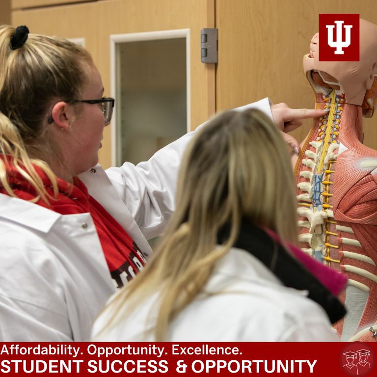 Two women in white coats point at a skeleton model
