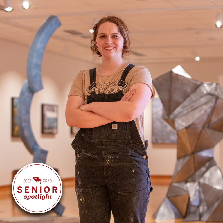A woman in overalls stands in front of two large metal sculptures