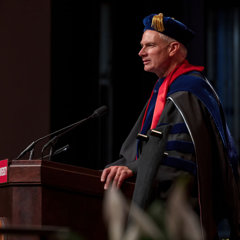 A man wearing academic robes speaks into a microphone