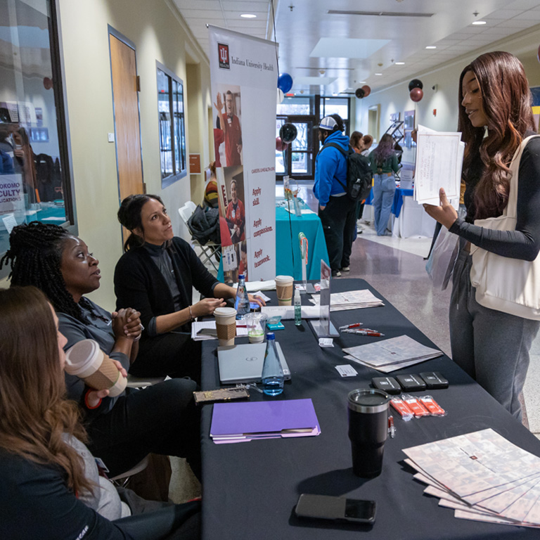 Three women seated behind a table talk to a woman standing in front of the table.
