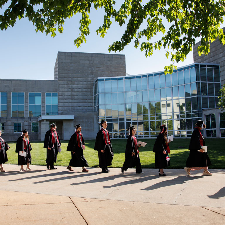 A line of graduates in caps and gowns walks by a large building