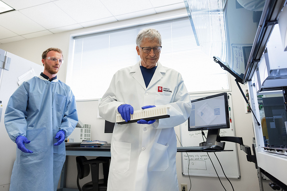 Bill Gates hold up a tray in a research lab, with an assistant in a blue lab coat in the background