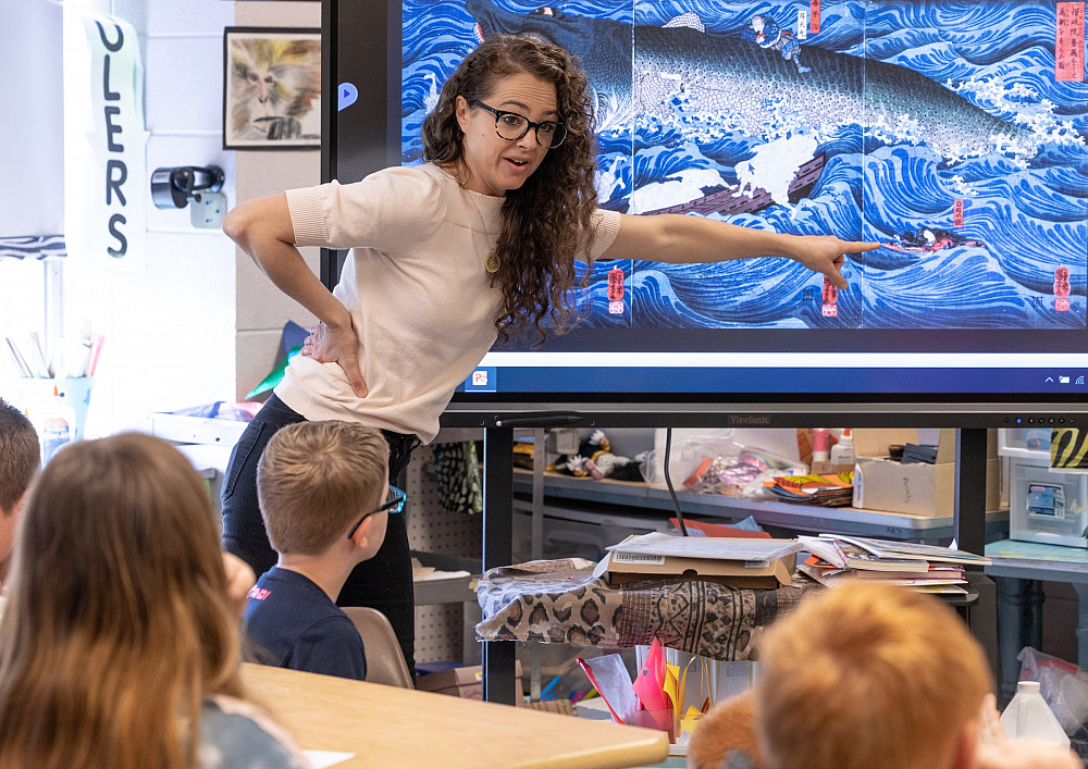 A woman in a classroom points to a screen that shows a beautiful blue art piece as students observe.