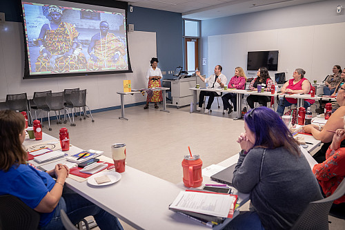 A woman stands at the front of a room presenting to teachers seated at tables around the perimeter