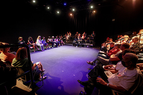 An audience sits around a black box theater stage bathed in purplish light.