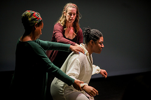Tow woman place their hands on the shoulders of a third woman is crouching slightly on a stage.