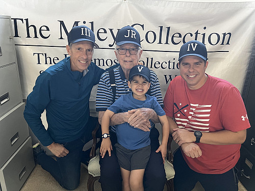 Four generations of John Mileys pose together in front of a banner that reads The Miley Collection: The Foremost Collection of Sports Aud...