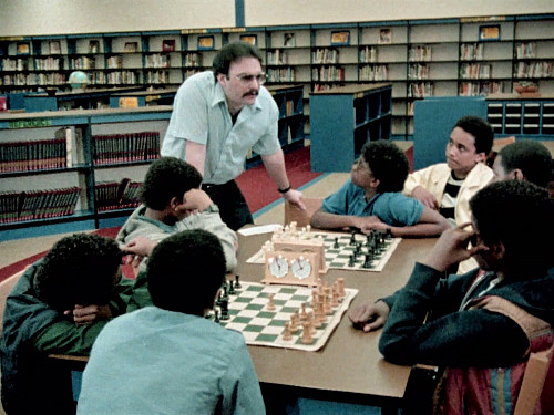 A man stands and speaks to young students sitting at a table covered in chess boards.