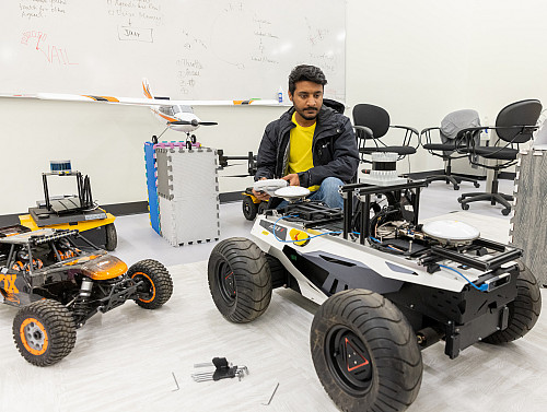 A man crouches in the middle of several small autonomous vehicles resembling remote controlled trucks and drones.