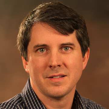 A man smiling in front of a brown background.