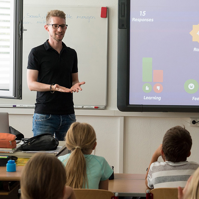 A male teacher stands in front of a classroom filled with students.
