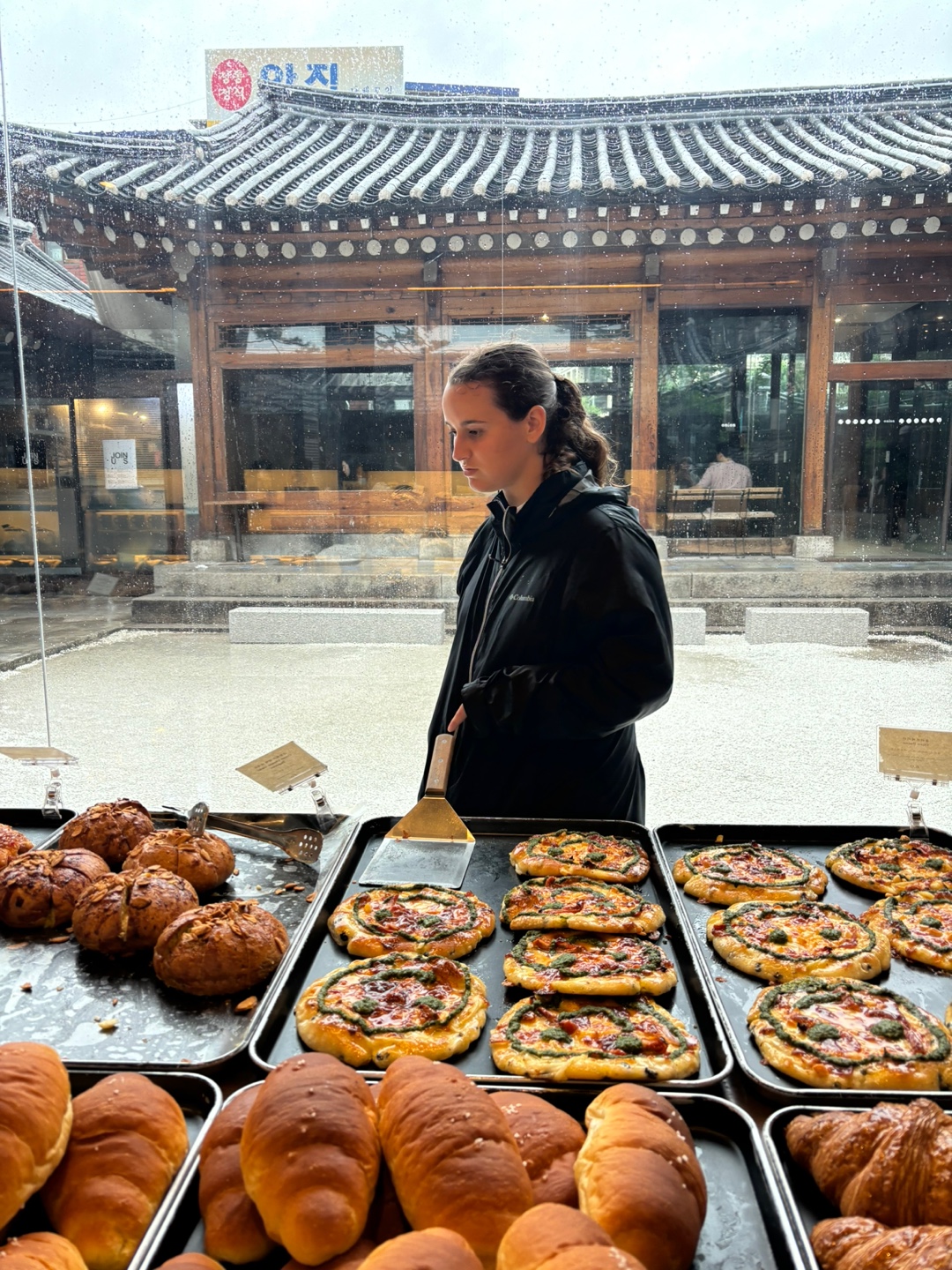 View from inside a South Korean bakery, looking out from the street, a woman stands in profile view, with a pagoda in the background
