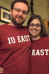 A couple arm in arm, smiling while wearing matching red IU East t-shirts in a living room.