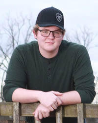 Corbin Frye in a baseball cap and glasses, leaning on a wooden railing in front of bare trees