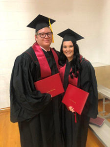 Couple stands side by side in their graduation cap, gown, and stoles, holding their diplomas