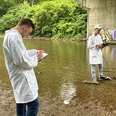 Two students in lab coats standing along the Whitewater River, testing the water quality and taking notes on a clipboard