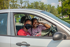Two people sitting in a car looking at the camera. The passenger is wearing a face mask and the driver is smiling