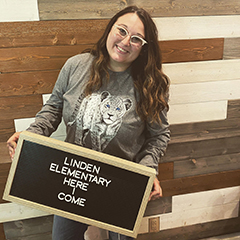 Aubrey Stevenson smiling while wearing a gray shirt featuring a white leopard and holding a sign that says Linden Elementary Here I Come.