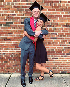 Graduating couple wearing their mortarboards and stoles, hugging in front of a brick wall