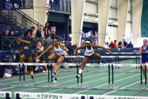 Sheliece Watkins and Kevaray Gillette leaping over hurdles at an indoor track and field meet