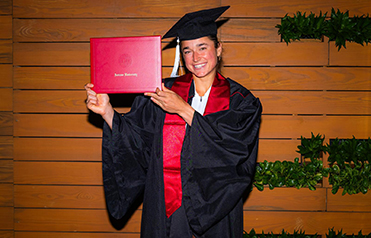 Dolehide smiling in her cap, gown and stole, holding up her red IU degree case