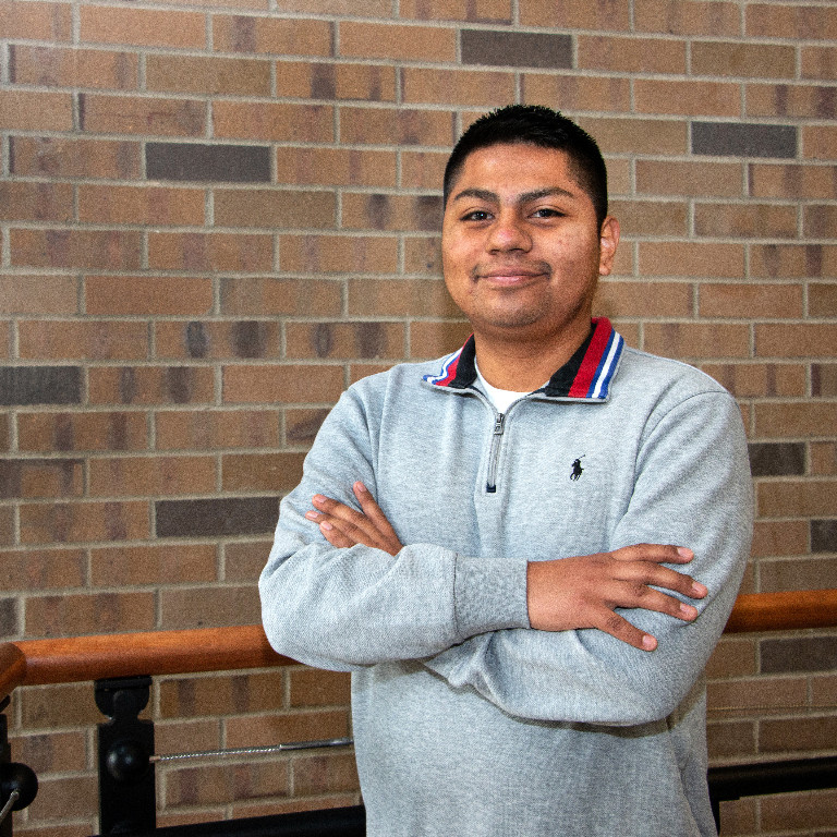 Eric Mejia smiling in front of a brick wall and railing with his arms casually crossed.