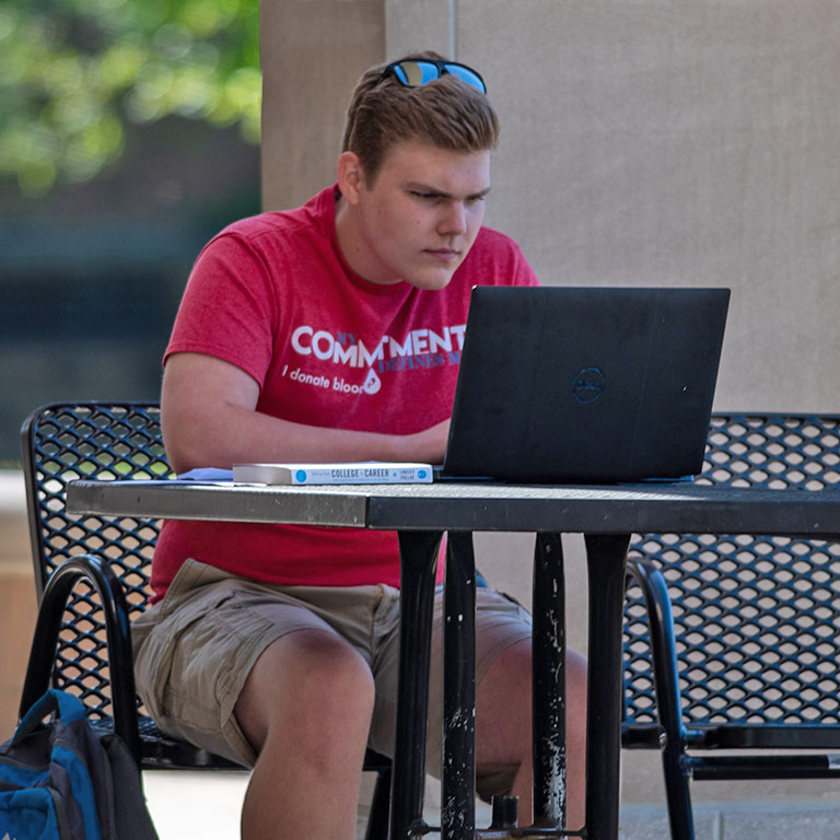 Student completing coursework online, seated outdoors and alone while practicing social distancing.