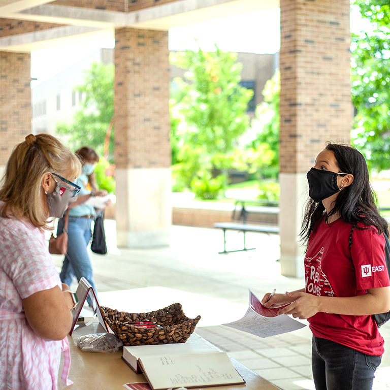 Masked staff member talks with a masked-up student on campus.