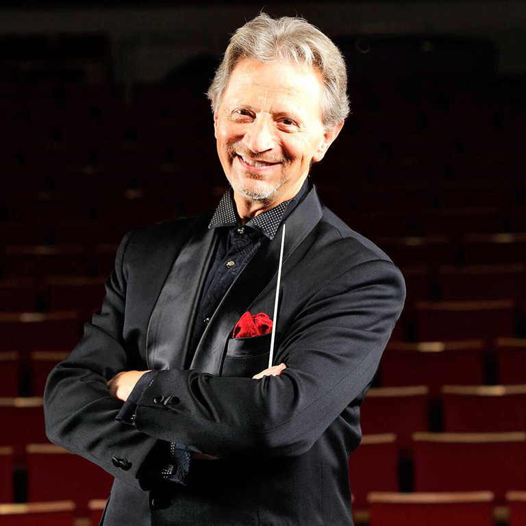 Jack Everly smiling in front of auditorium seating, his arms casually crossed and a conductor's baton in his hand.