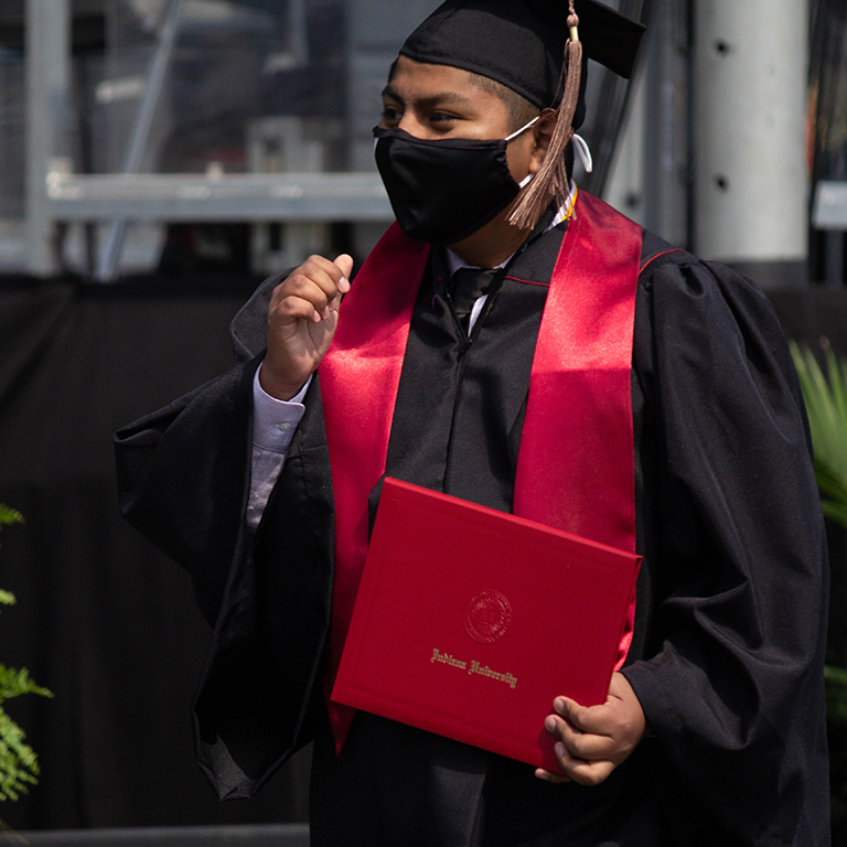 Eric Mejia dressed in a graduation cap, gown, and stole, holding his diploma cover at Commencement