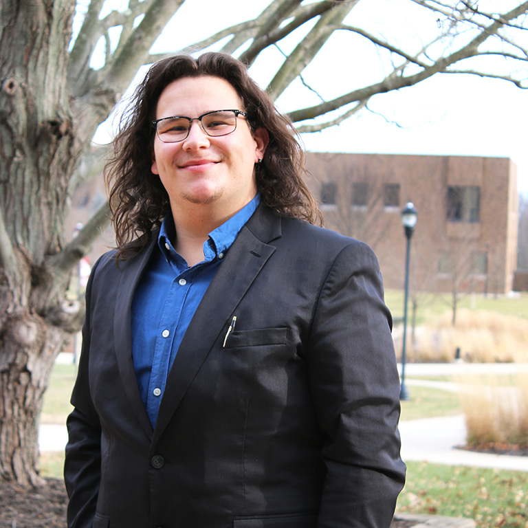 Alex Hakes smiling in front of a tree and campus walkway