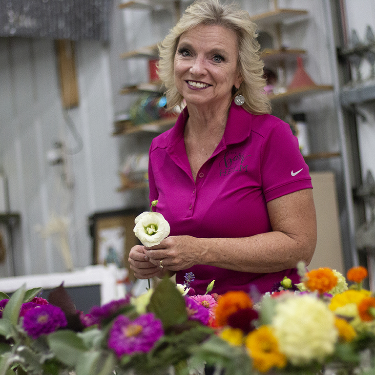 Julie Frame smiling and holding a flower with bright flowers in the foreground.