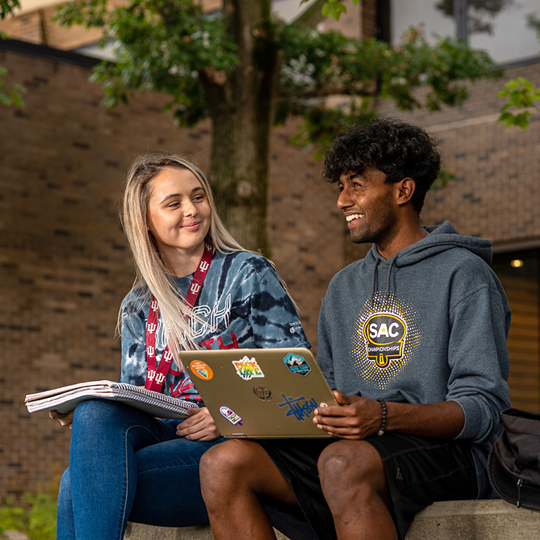 Two students smiling and sitting together on the patio of Whitewater Hall. One has a notebook and the other a laptop.