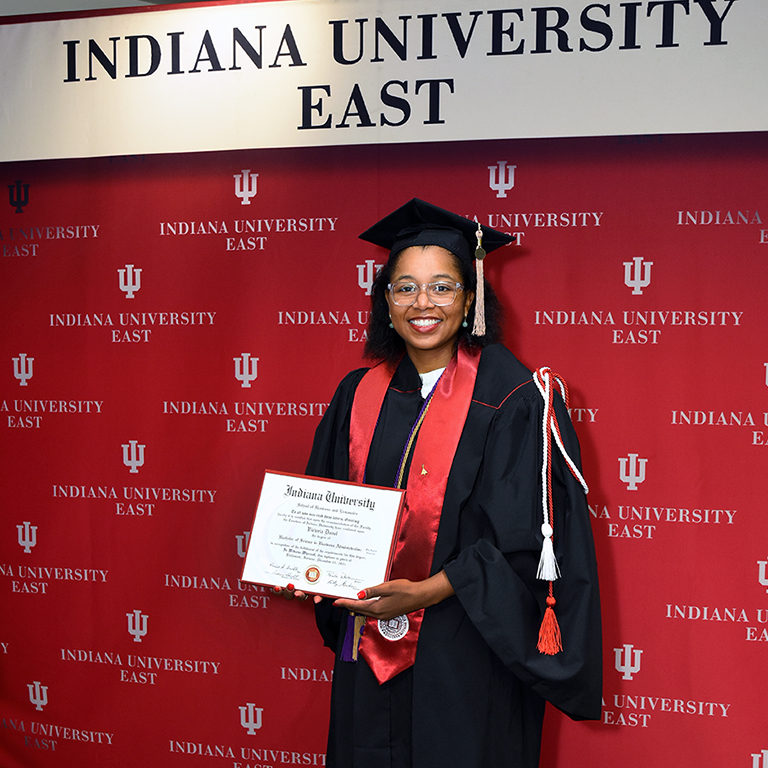 Vicky Duval standing with her diploma in front of the IU East media backdrop